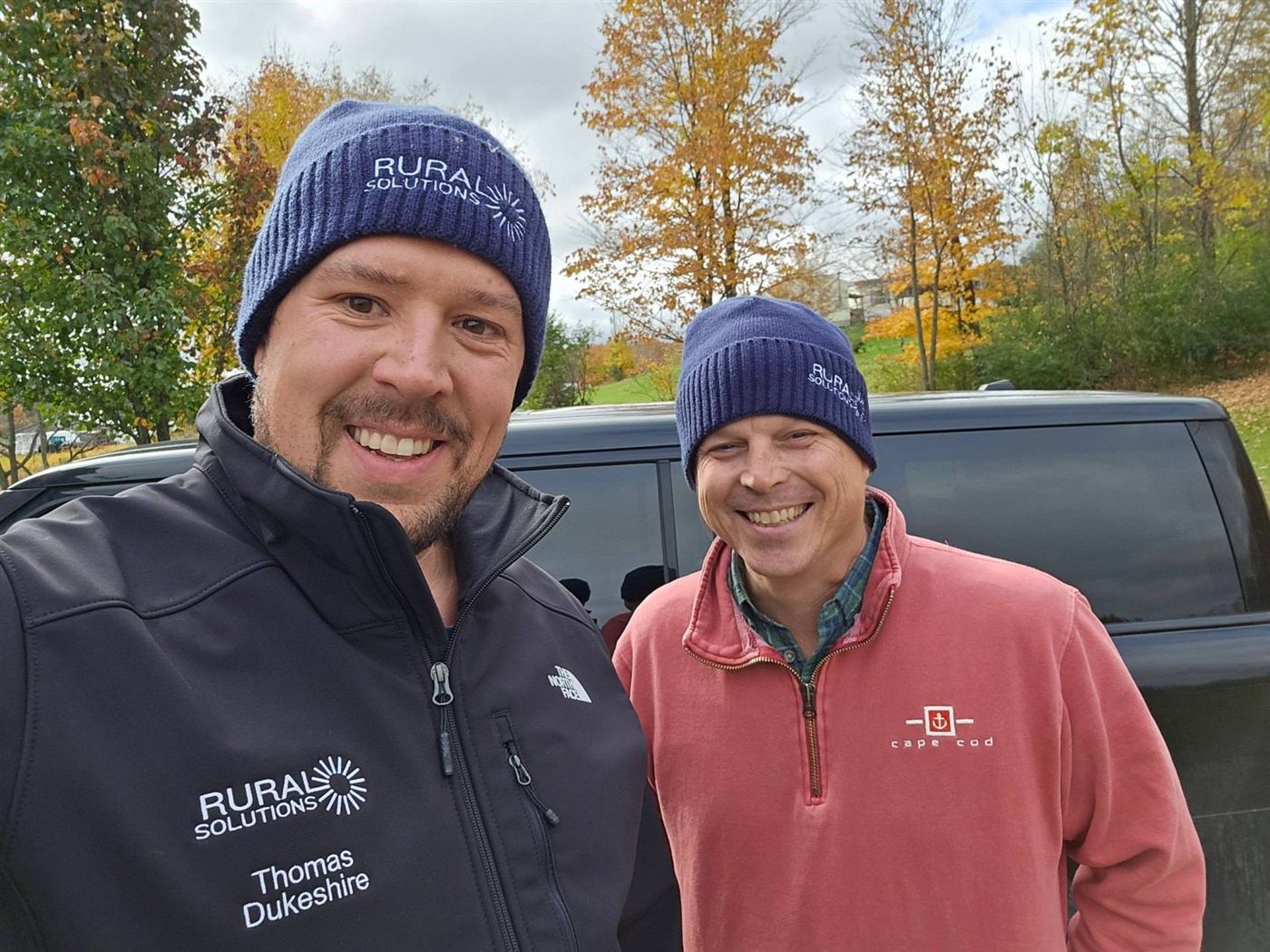 Thomas Dukeshire and Allen Nichols of Rural Solutions, wearing Rural Solutions hats, smiling beside a vehicle on an autumn day in Vermont